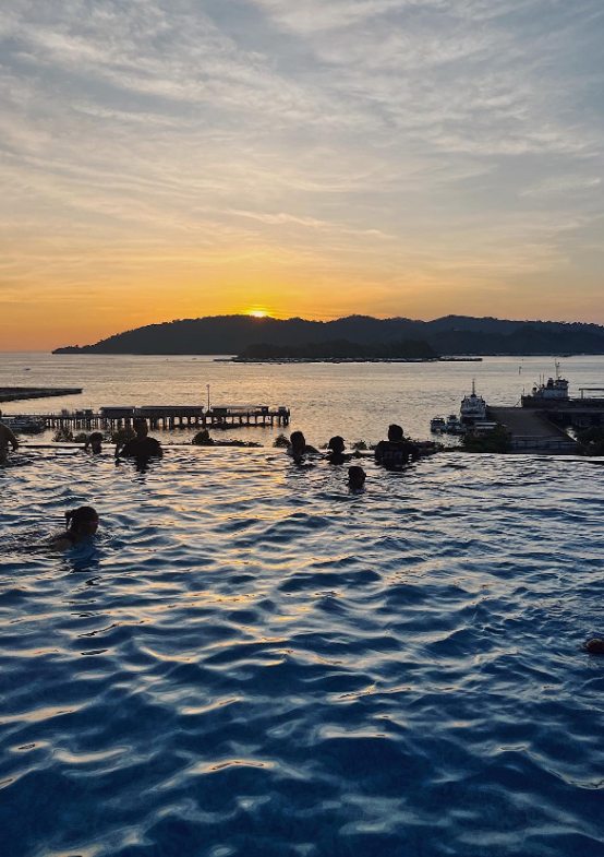 遊客在泳池中欣賞沙巴夕陽，遠方可見海灣、碼頭與島嶼。