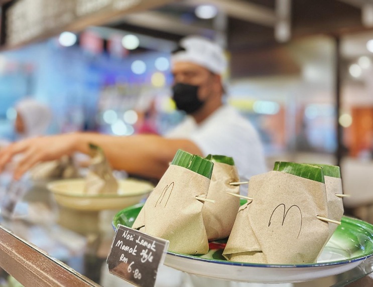 攤位上陳列以香蕉葉和牛皮紙包裹的椰漿飯，後方店員正在備餐。