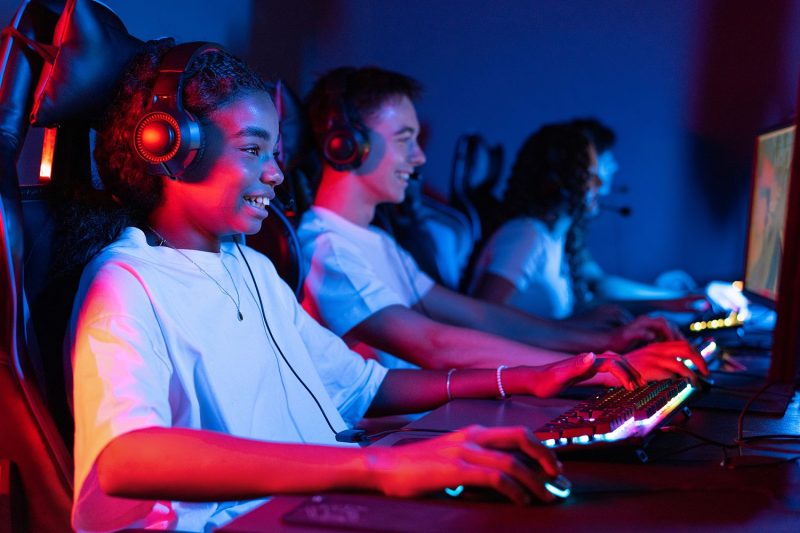Teens playing in video game club Group of multiracial teens in headsets playing video games in video game club with blue and red illumination. Keyboard and mouse with illumination